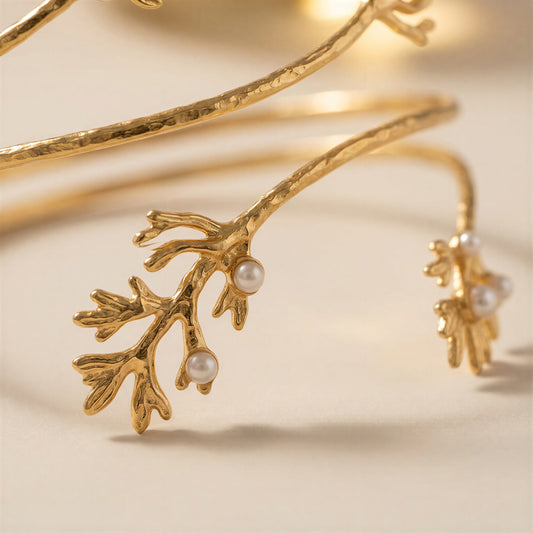 Gold branch-shaped bangle bracelet with small white pearl accents, photographed on a warm cream background.