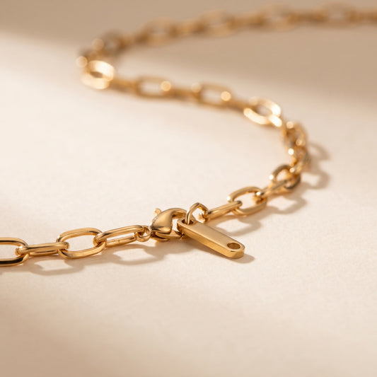 A close-up of a gold chain necklace with a lobster clasp and a small rectangular tag, photographed on a warm beige surface with soft shadows.