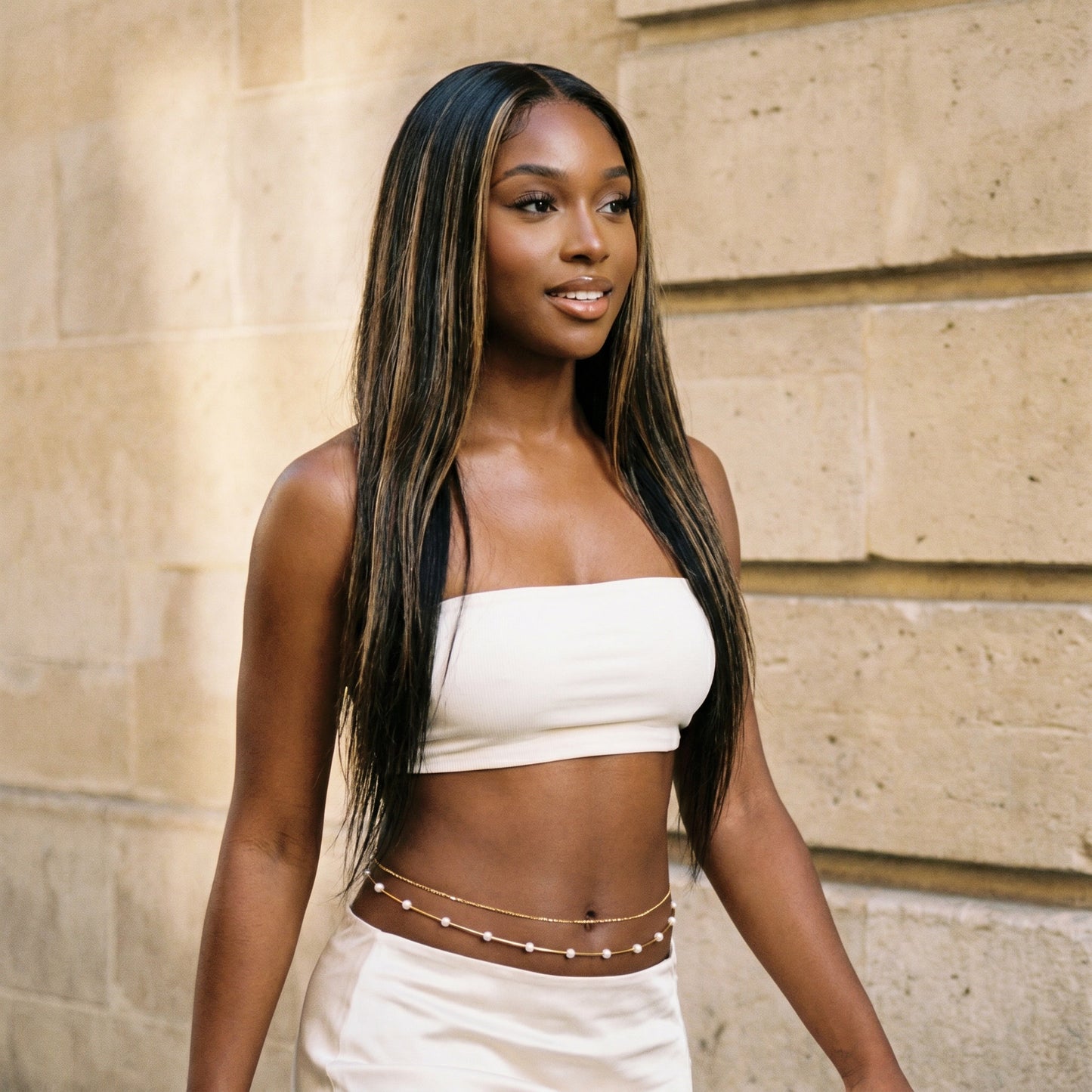 A woman with long, straight black hair with blonde highlights is wearing a white strapless tube top and matching white bottoms with a gold chain belt, standing against a beige stone wall.