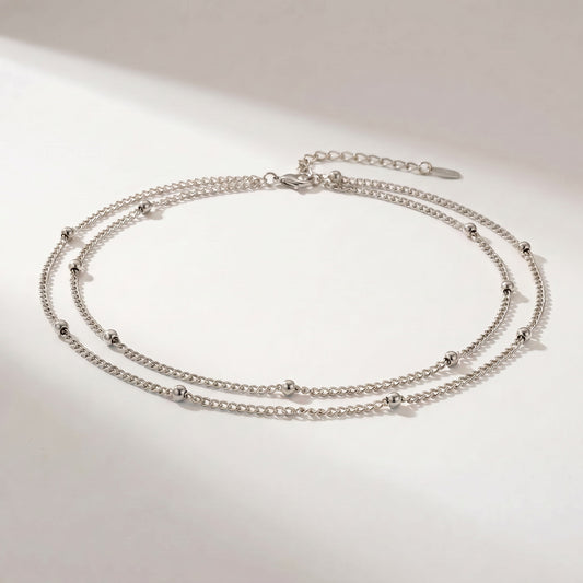 A silver double-layered chain anklet with small bead accents, displayed on a clean white surface with soft shadows.