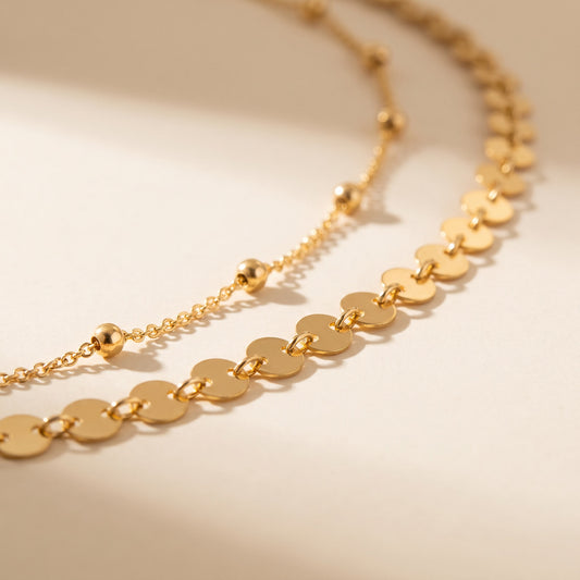 A close-up of a gold-toned necklace with flat circular disc links and a few small bead accents, photographed on a warm off-white background with soft shadows.