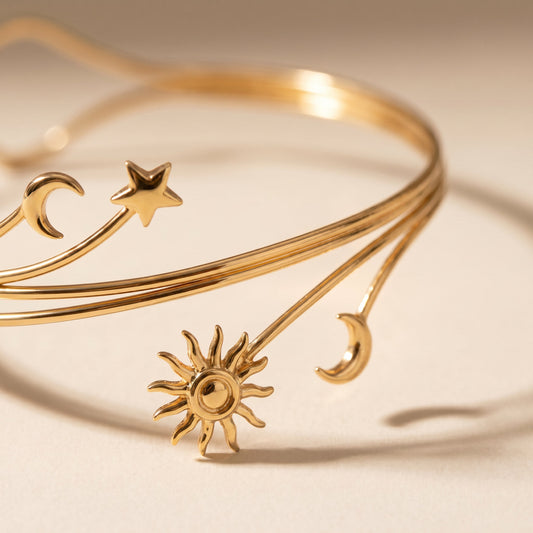 A delicate gold-toned bangle bracelet with celestial motifs, including a sun, crescent moons, and stars, displayed on a warm beige background.