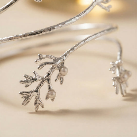 A close-up of a silver bangle bracelet designed to look like a branch with small pearl-like beads, photographed on a soft beige background.