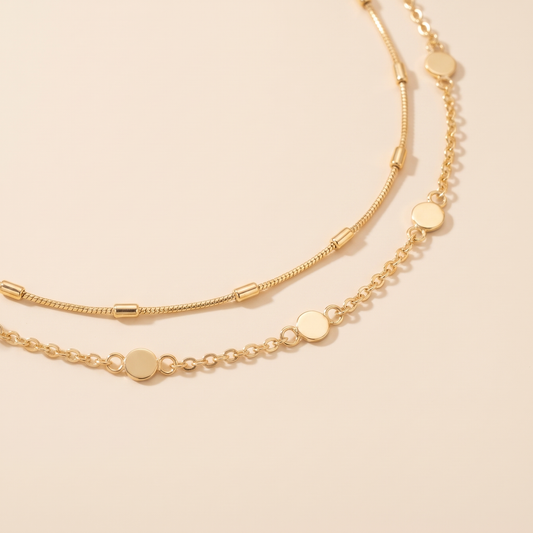 A delicate gold chain necklace with flat circular disc charms, displayed on a warm beige surface with a soft, warm-toned shadow.