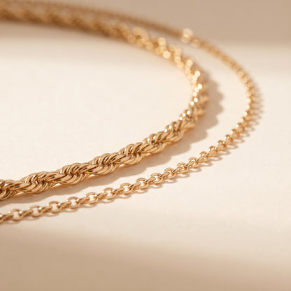A close-up of a gold chain necklace draped in two parallel loops on a warm beige surface with soft shadows.
