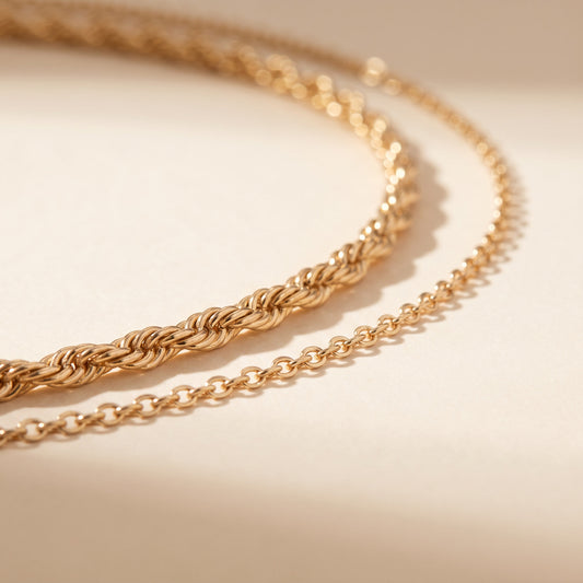 A close-up of a gold chain necklace draped in two parallel loops on a warm beige surface with soft shadows.