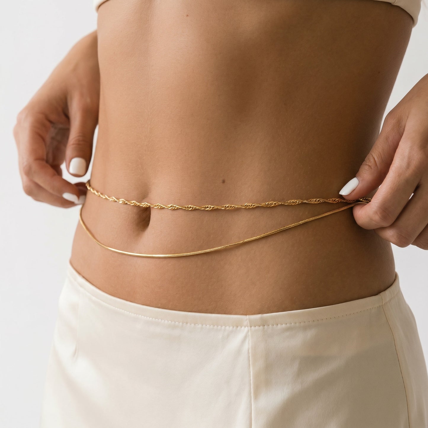 A close-up of a model's midsection wearing two delicate gold chain waist accessories, one twisted and one smooth, paired with cream-colored bottoms.