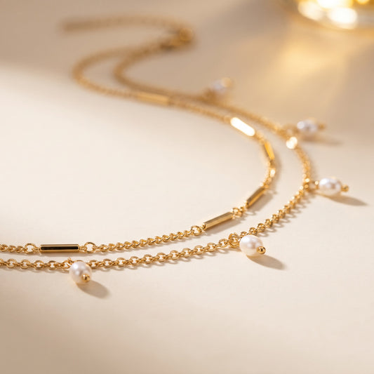 A delicate gold double-strand chain necklace with small white pearl beads and polished gold bar accents, displayed on a warm beige surface with soft, warm-toned lighting.