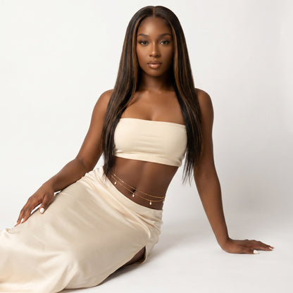 A woman with long, straight dark brown hair models a matching cream-colored two-piece set consisting of a strapless crop top and a high-waisted wrap skirt, accessorized with delicate gold chain details. She is posed on a seamless light gray studio background.