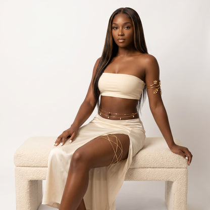 A woman with long dark hair sits on a cream boucle bench wearing a matching cream two-piece set with a strapless bandeau top and a high-slit skirt, accented with gold chain details on her waist, hips, and upper thigh.