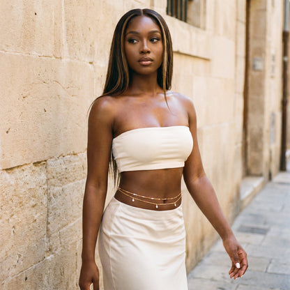 A woman stands outdoors against a beige stone wall wearing a matching off-white strapless tube top and high-waisted skirt, with thin gold chain waist accessories.