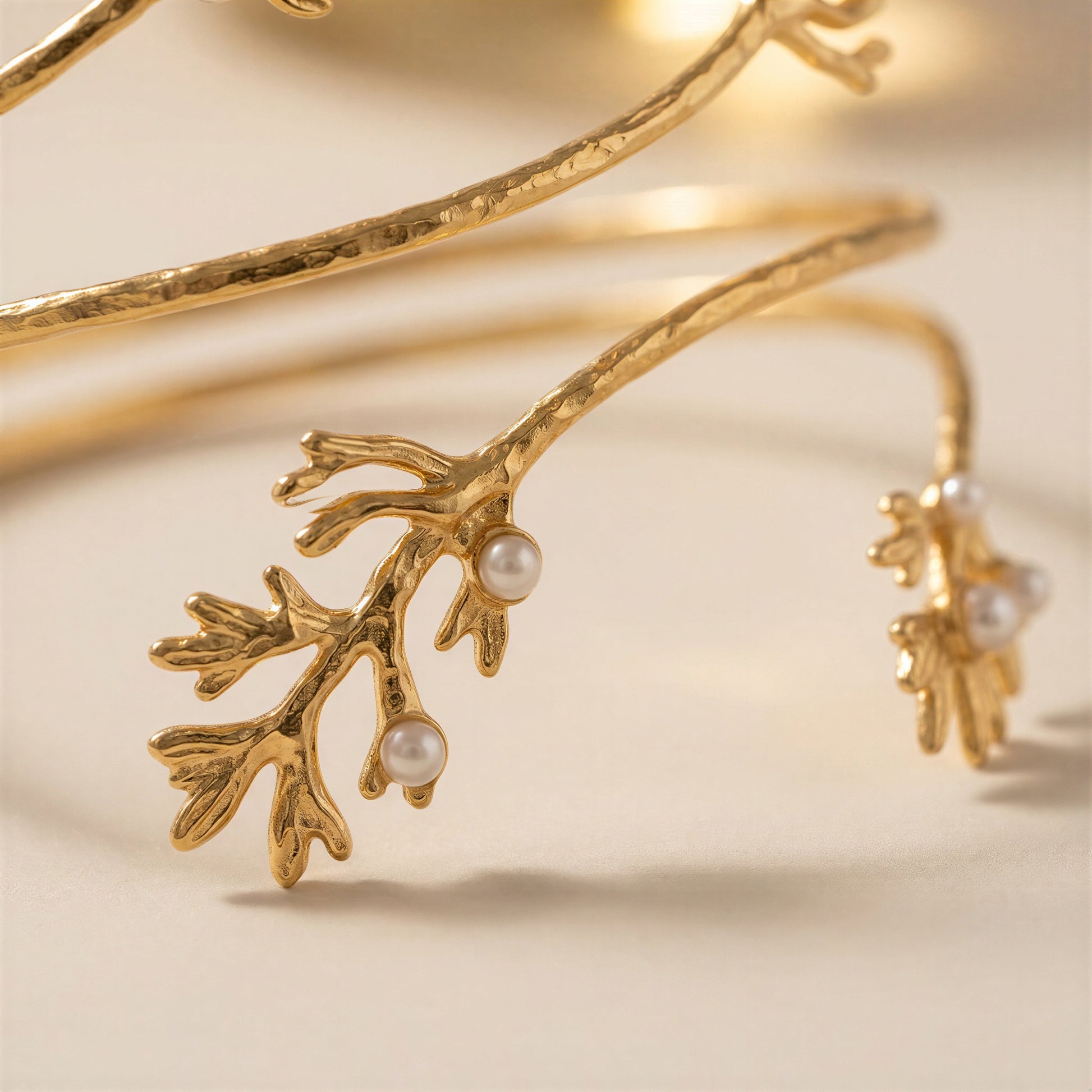 Gold branch-shaped bangle bracelet with small white pearl accents, photographed on a warm cream background.