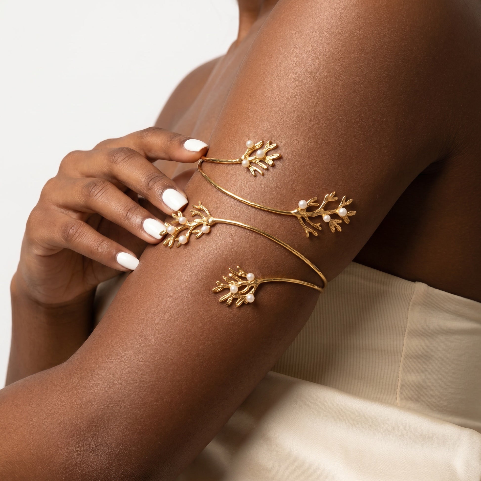 A close-up of a model's arm wearing a delicate gold open cuff bracelet decorated with three leafy branches and small pearl-like beads, against a soft light background.