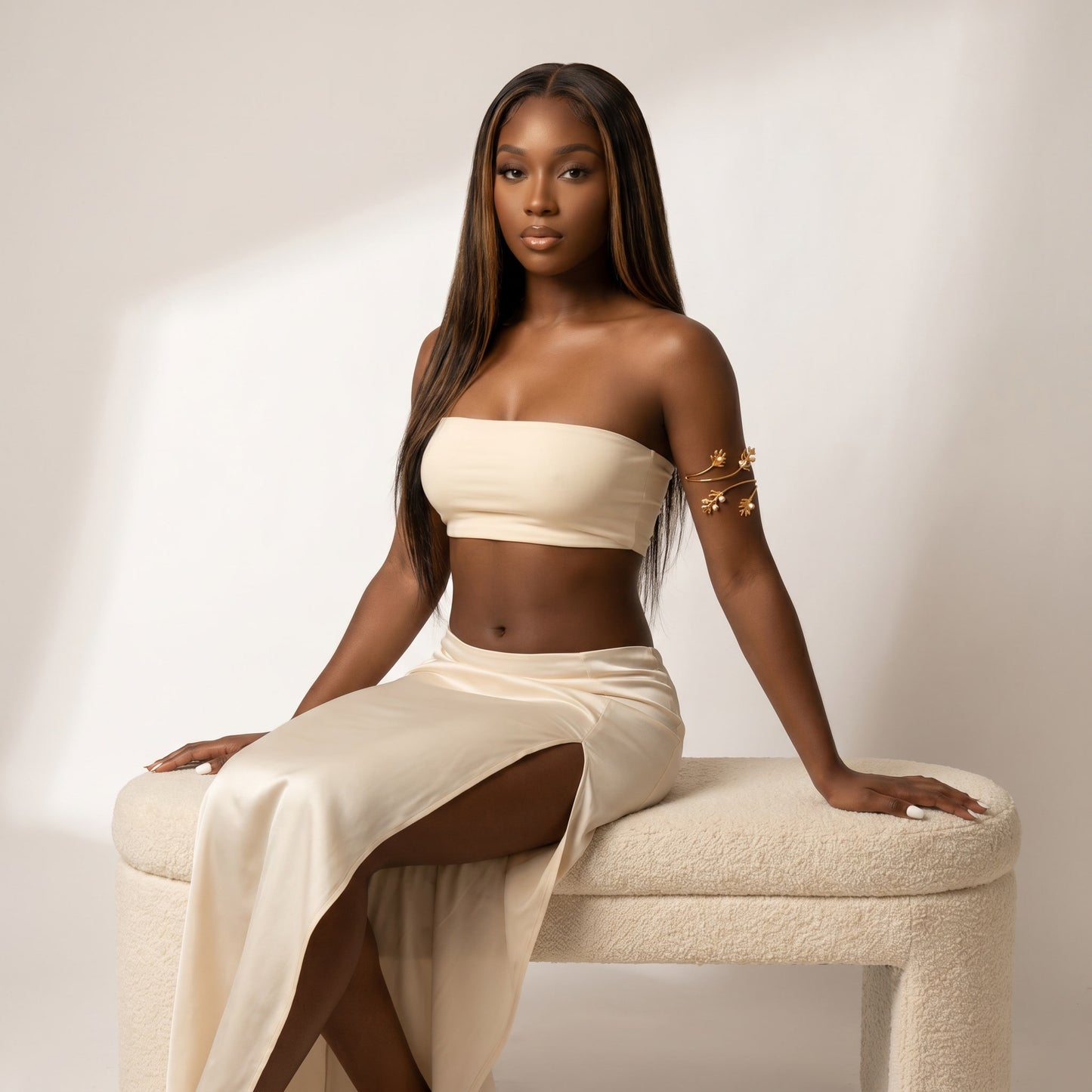 A woman with long dark hair sits on a cream boucle bench wearing a matching cream two-piece set consisting of a strapless tube top and a high-slit skirt. She is wearing a gold armband bracelet.