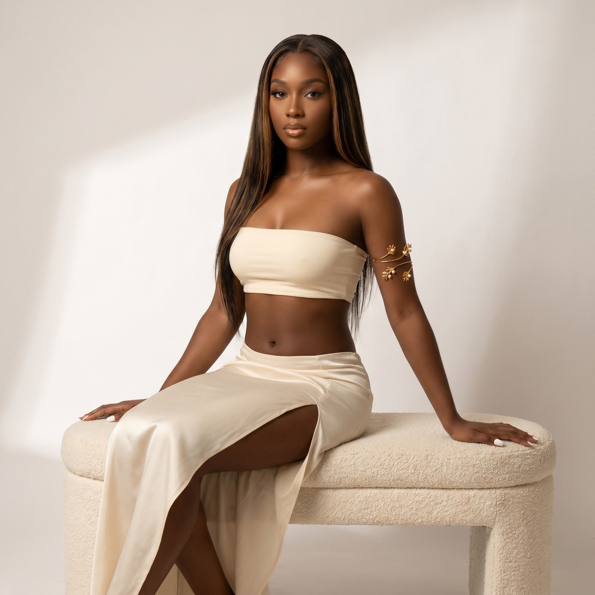 A woman with long dark hair sits on a cream boucle bench wearing a matching cream two-piece set consisting of a strapless tube top and a high-slit skirt. She is wearing a gold armband bracelet.