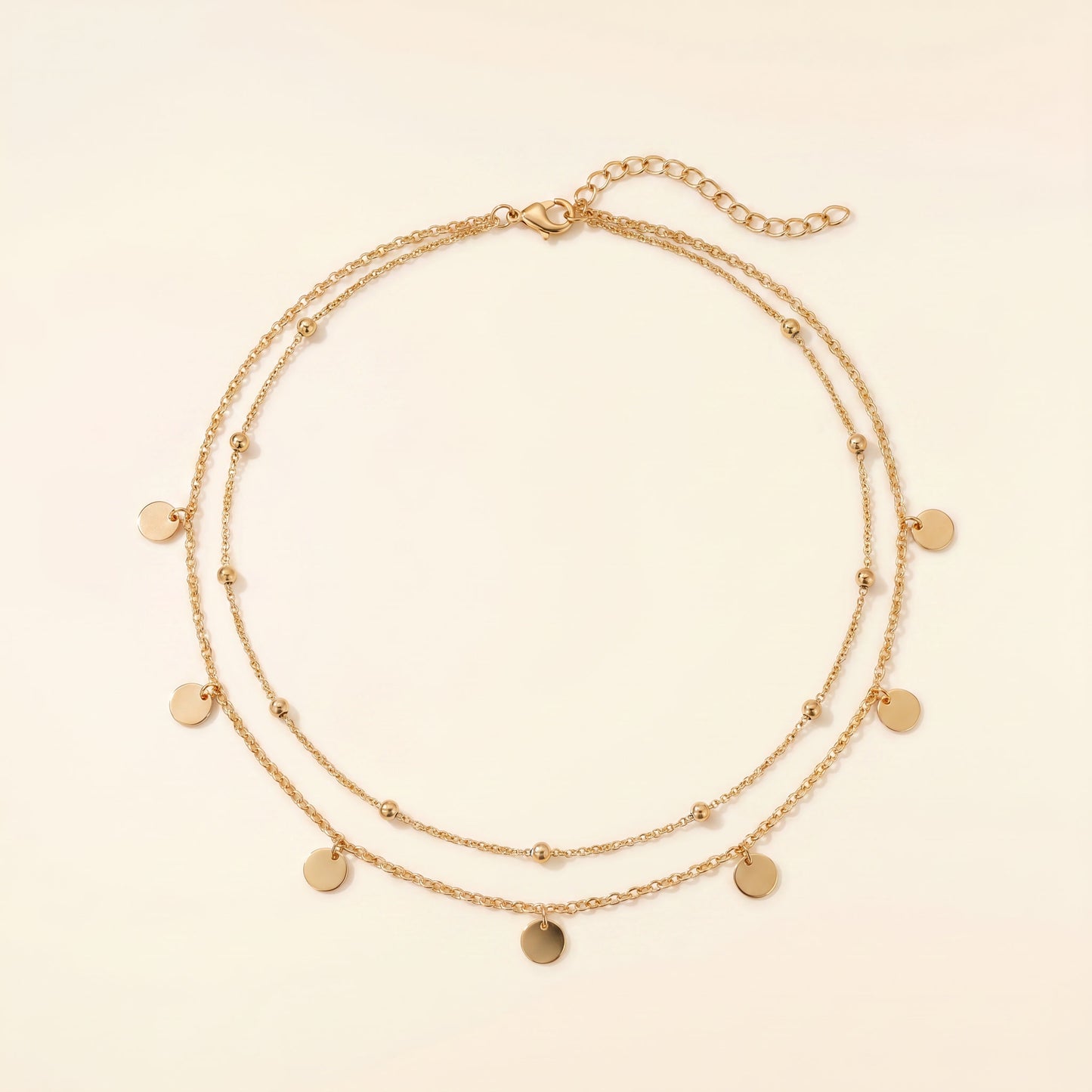 A double-strand gold chain necklace with small flat round disc charms and tiny bead accents, displayed on a light cream background.