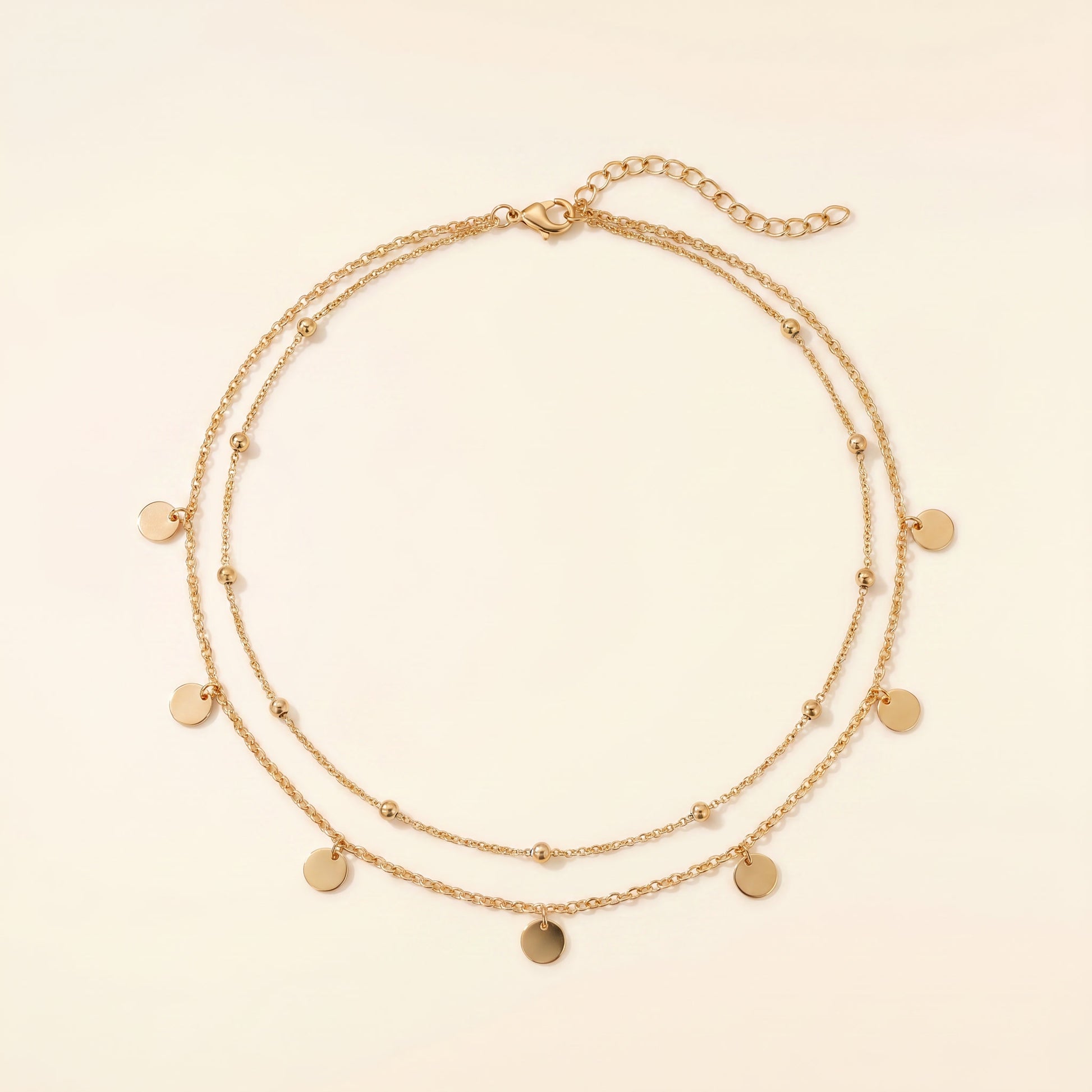 A double-strand gold chain necklace with small flat round disc charms and tiny bead accents, displayed on a light cream background.