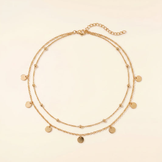 A double-strand gold chain necklace with small flat round disc charms and tiny bead accents, displayed on a light cream background.