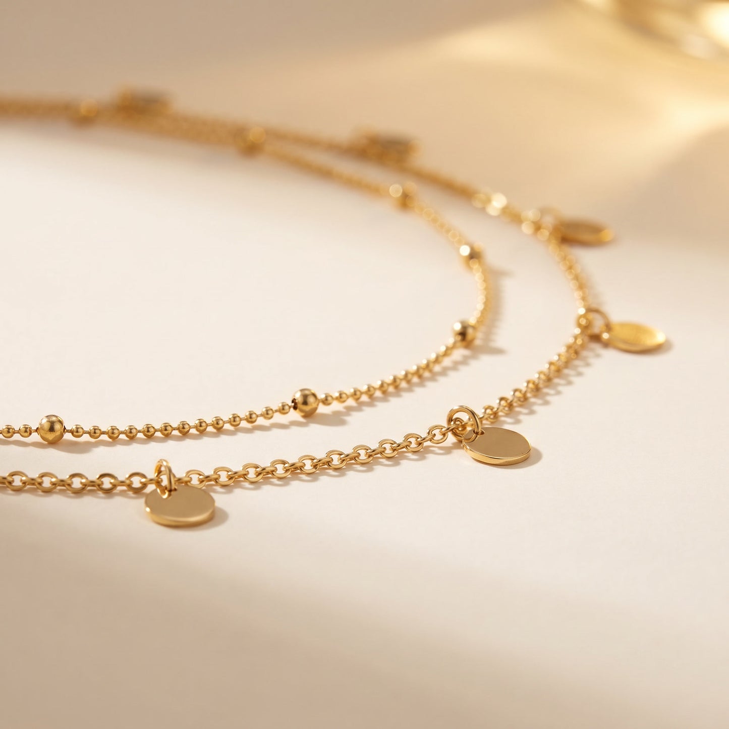 A delicate gold double-layer necklace with small disc charms and bead details, displayed on a warm beige background.