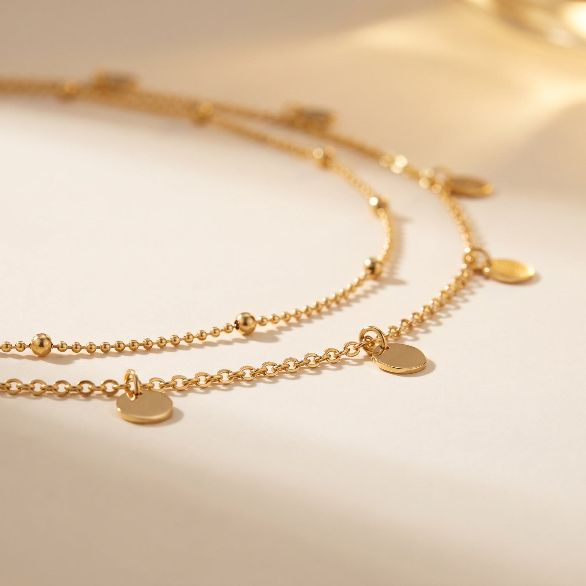 A delicate gold double-layer necklace with small disc charms and bead details, displayed on a warm beige background.