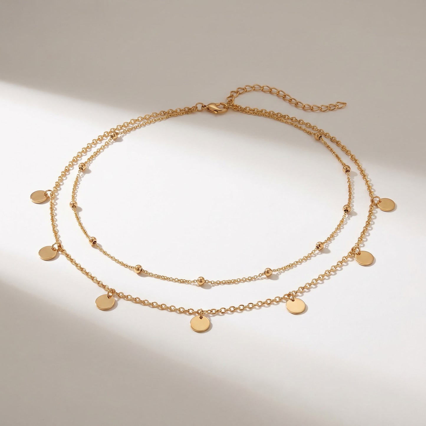 A double-layered gold chain necklace with small flat round disc charms and tiny bead accents, displayed on a light beige background with soft diagonal shadows.