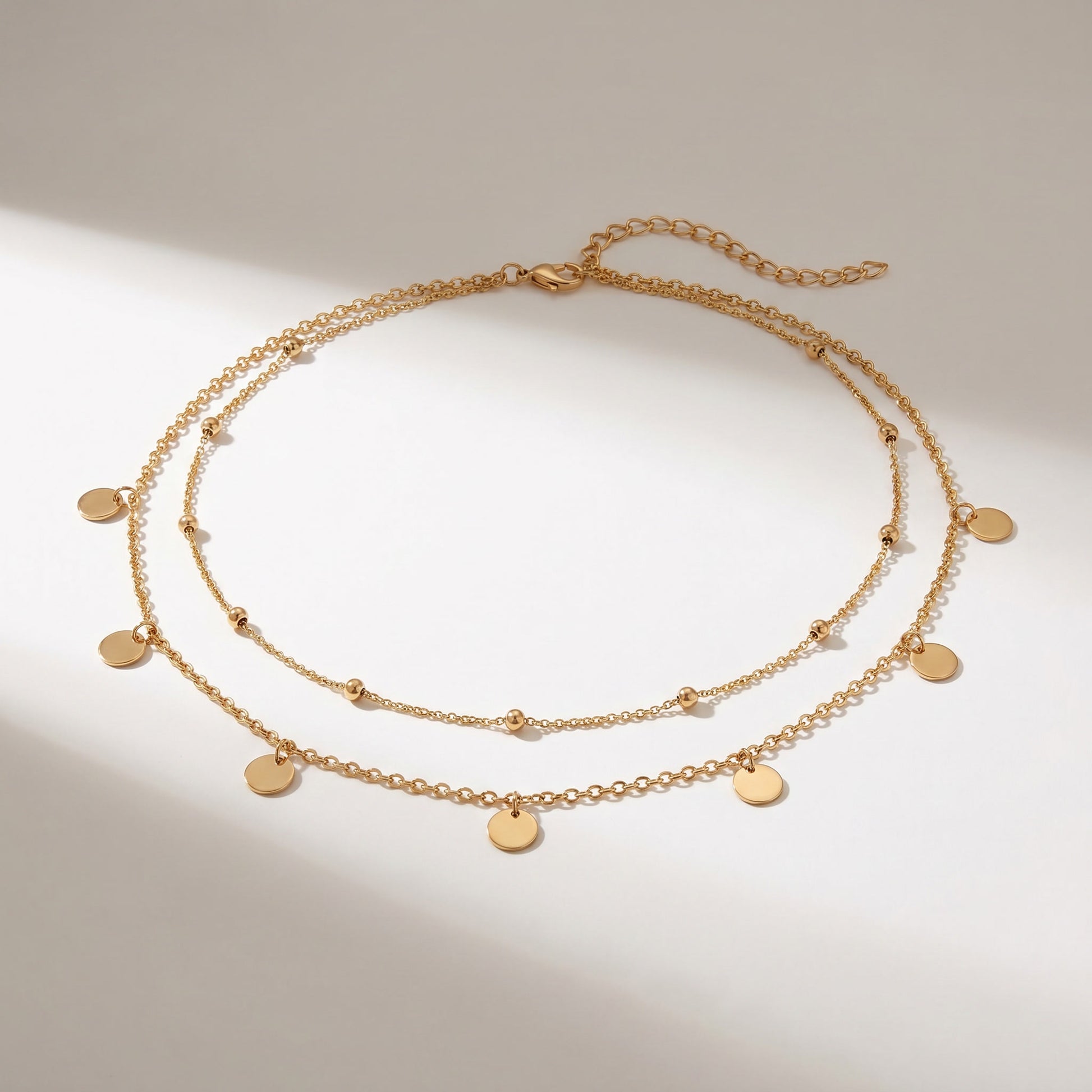 A double-layered gold chain necklace with small flat round disc charms and tiny bead accents, displayed on a light beige background with soft diagonal shadows.