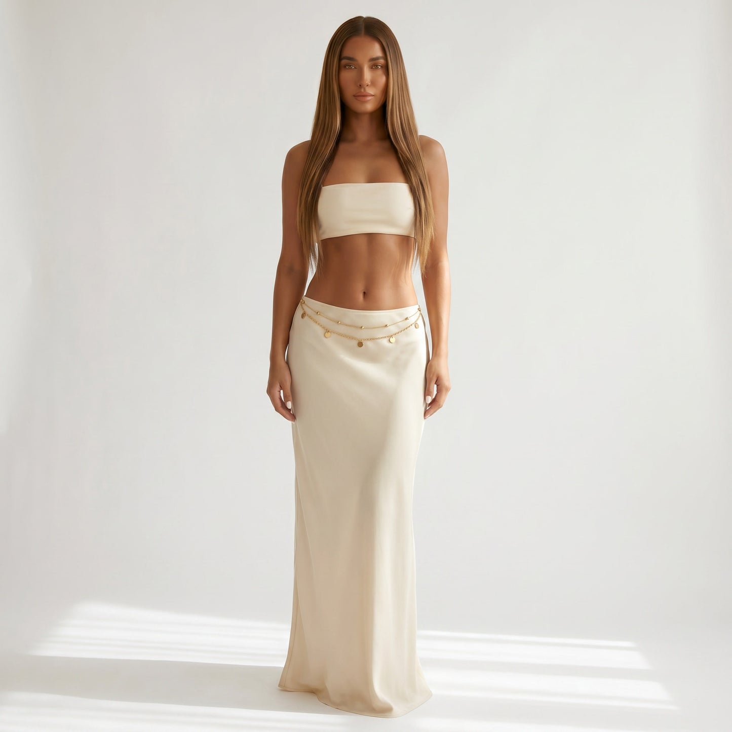 A woman stands against a bright white studio background wearing a matching two-piece outfit: a strapless bandeau-style top and a long skirt in a warm cream or off-white tone, with a thin chain detail at the waistband.