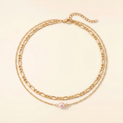 A double-layered gold chain necklace featuring a classic Figaro chain and a delicate fine chain with a single pink pearl pendant, displayed on a light cream background.