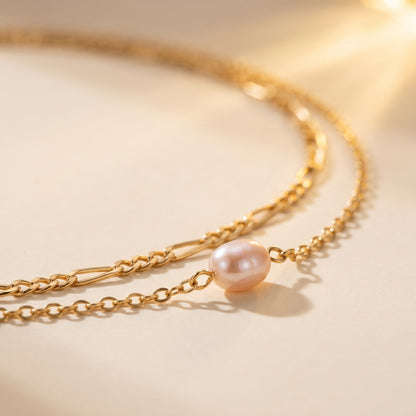 A delicate gold chain necklace featuring a single lustrous pearl pendant, photographed on a warm beige background with soft sunlight and shadows.