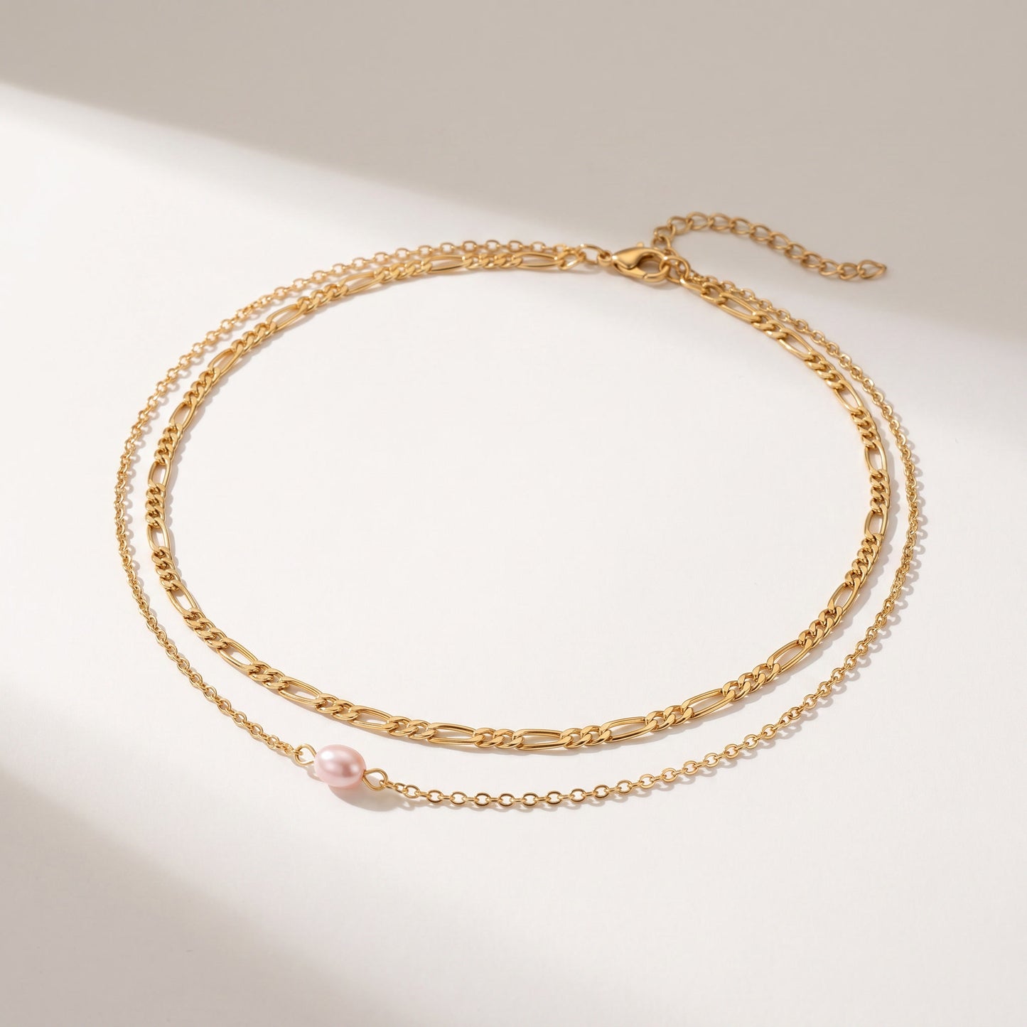 A delicate gold-toned double-layer necklace featuring a small pink freshwater pearl accent, displayed on a clean white surface with soft shadows.