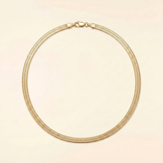 A gold-toned metal snake chain necklace with a lobster clasp, displayed in a circular shape against a solid pale pink background.