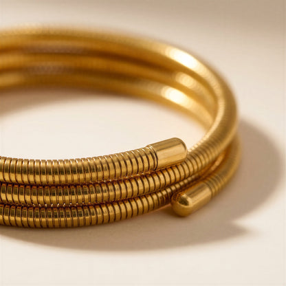A close-up of a gold-toned coiled bangle bracelet with a textured, ribbed outer surface and smooth cylindrical end caps, photographed on a warm off-white background.