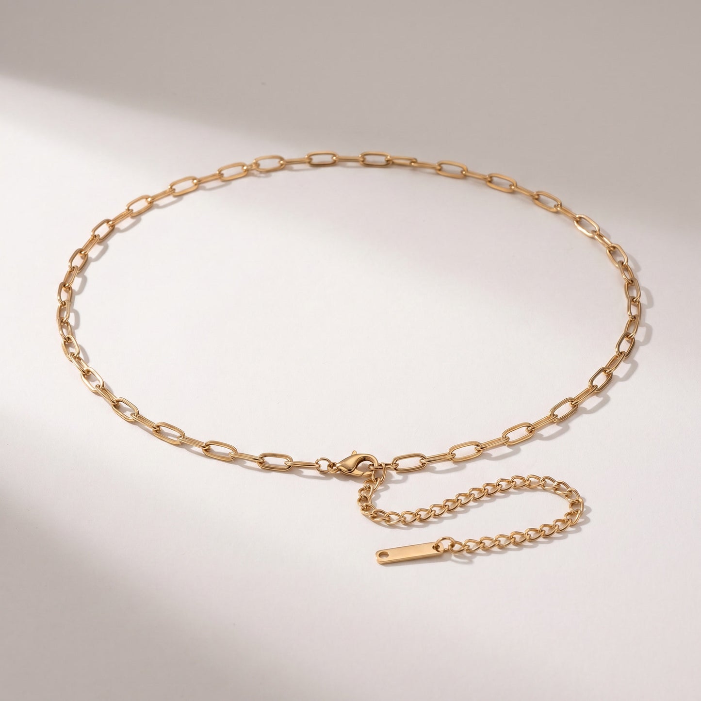 A gold chain belt with a flat, polished finish, featuring a spring-ring clasp and an adjustable extender chain with a small rectangular tag, displayed on a light gray background.