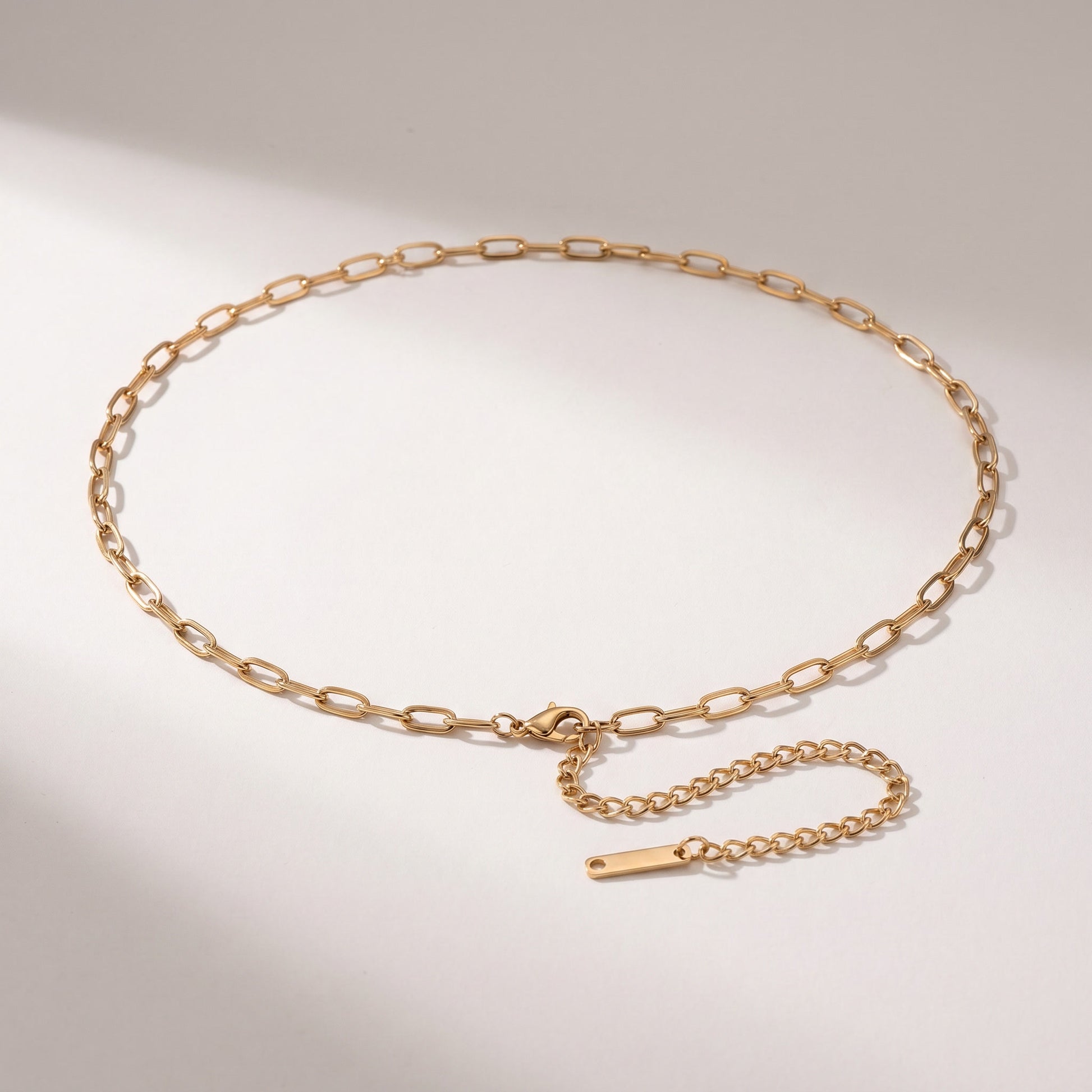 A gold chain belt with a flat, polished finish, featuring a spring-ring clasp and an adjustable extender chain with a small rectangular tag, displayed on a light gray background.