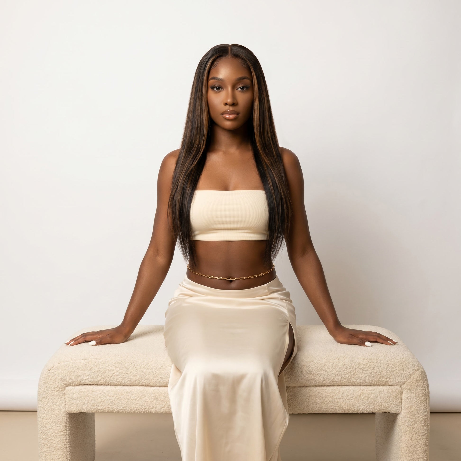A woman with long, straight dark hair sits on a cream boucle bench against a plain off-white wall, wearing a matching cream strapless tube top and high-waisted satin skirt.