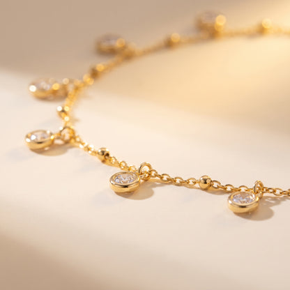 A close-up of a delicate gold chain necklace with small oval bezel-set clear stones, photographed on a warm cream background.