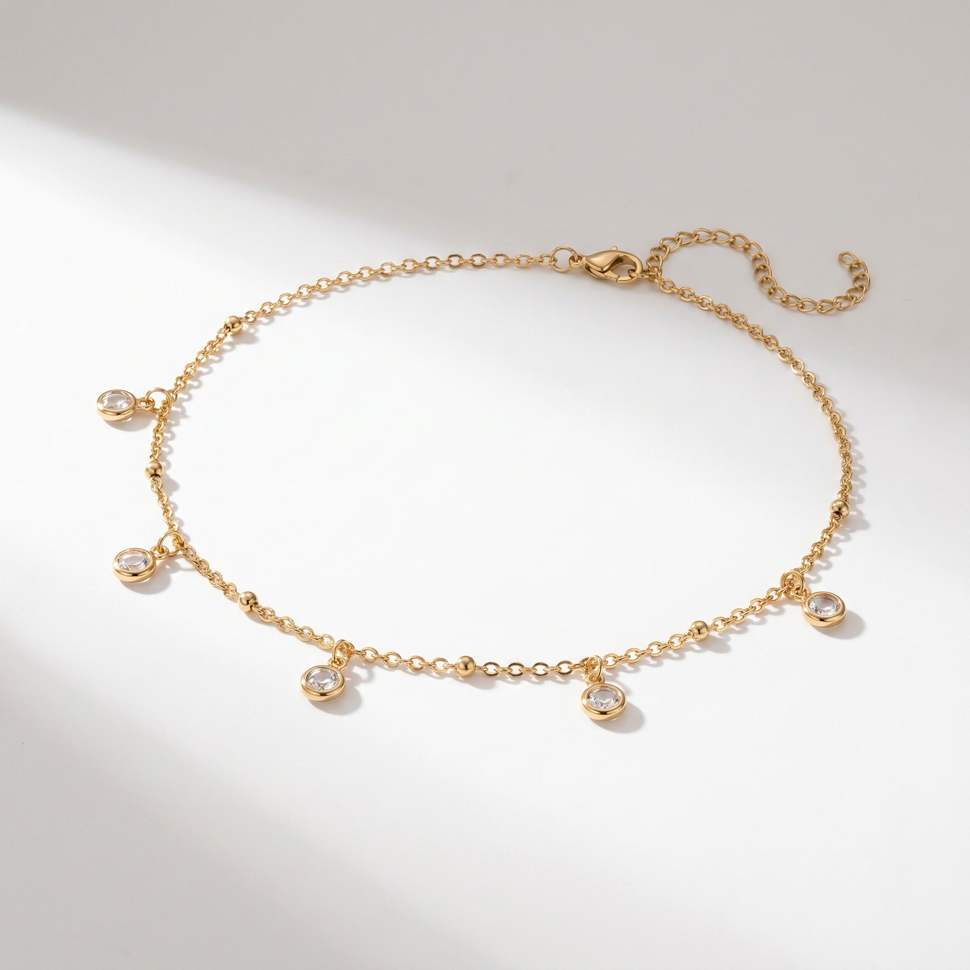 A delicate gold chain necklace with five small clear gemstone charms, displayed on a clean white background with soft shadows.