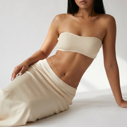 A woman wearing a cream-colored ribbed bandeau tube top and a matching cream skirt, accessorized with a delicate gold chain belt, posed against a clean white studio background.