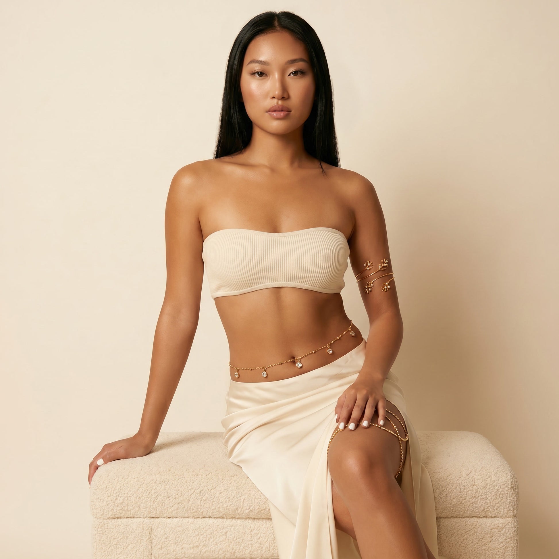 A woman models a matching beige ribbed bandeau top and satin skirt set, accessorized with gold chain bracelets and a chain belt, sitting on a textured cream boucle ottoman against a warm beige background.