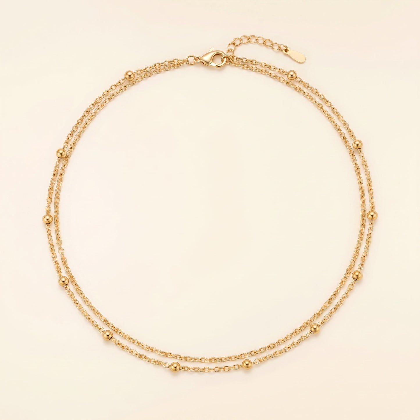 A delicate double-strand gold chain necklace with small bead accents, displayed on a light cream background.