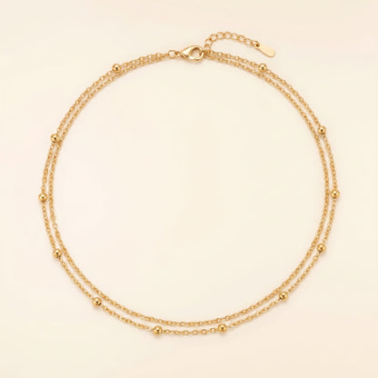 A delicate double-strand gold chain necklace with small bead accents, displayed on a light cream background.