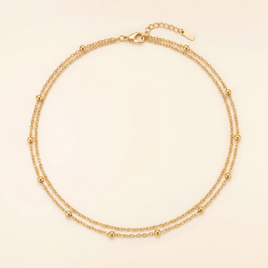 A delicate double-strand gold chain necklace with small bead accents, displayed on a light cream background.