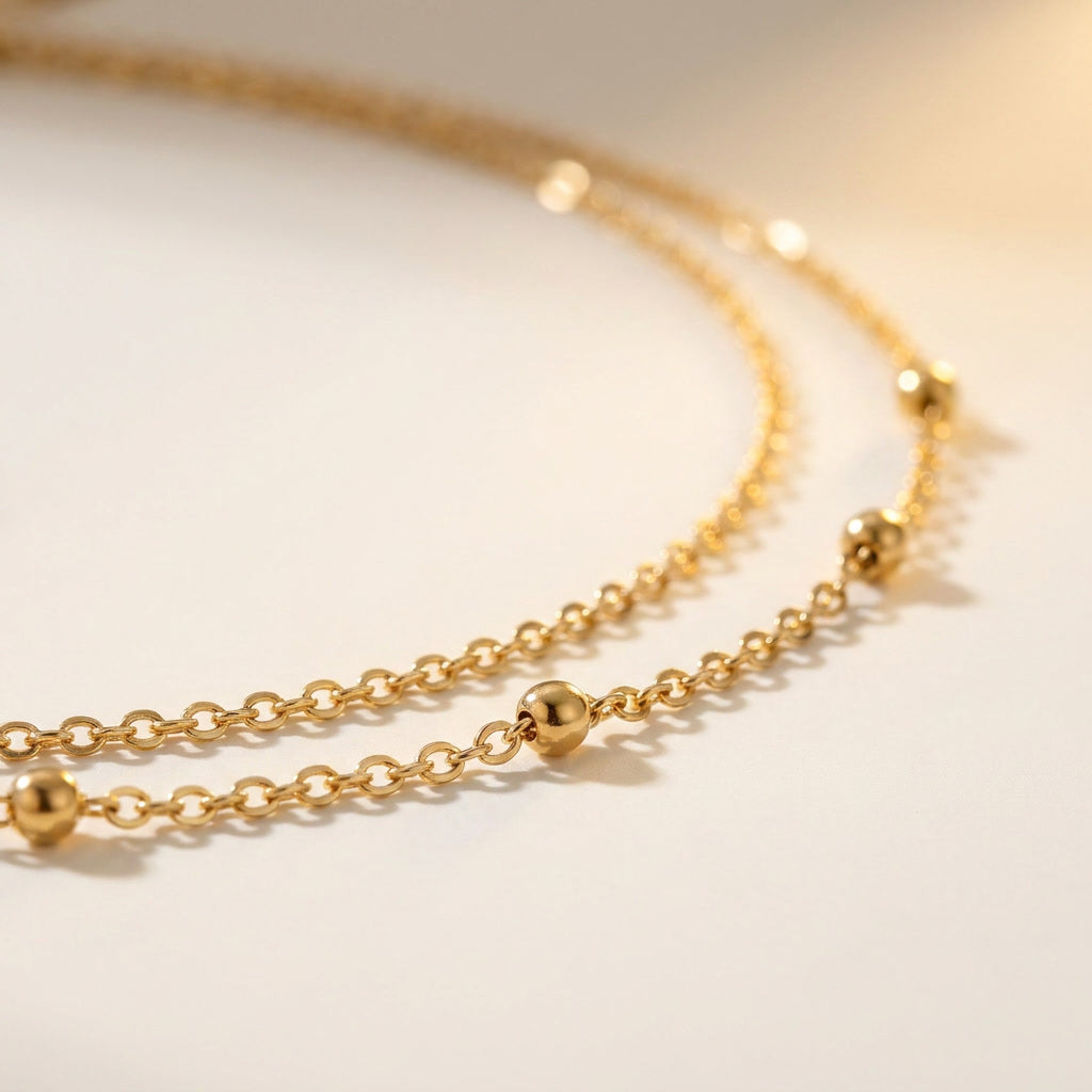 A close-up of a delicate gold chain necklace with small polished bead accents, photographed on a soft off-white background.