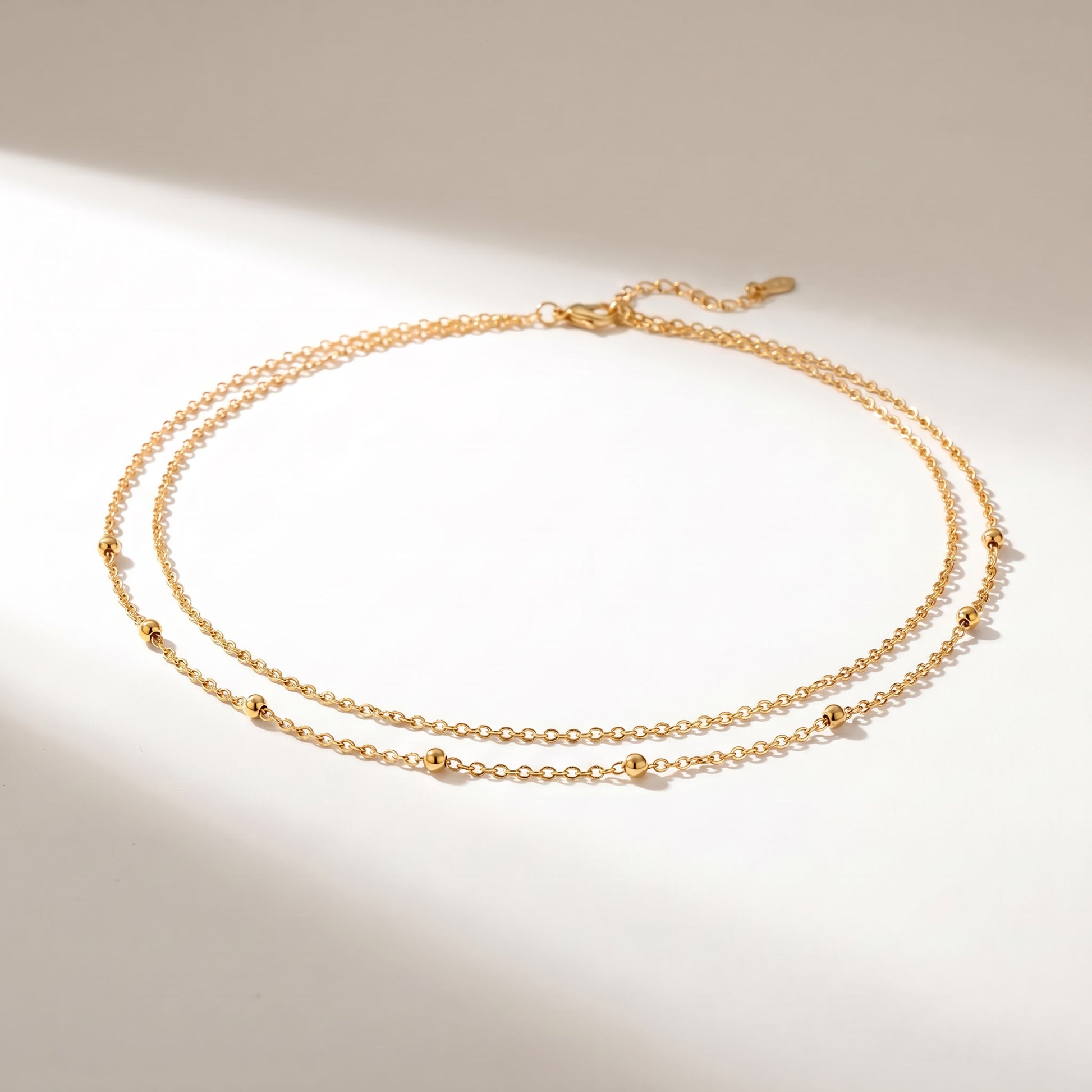 A delicate double-strand gold chain necklace with small spaced bead accents, displayed on a warm off-white surface with soft diagonal shadows.