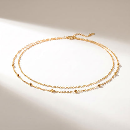 A delicate double-strand gold chain necklace with small spaced bead accents, displayed on a warm off-white surface with soft diagonal shadows.