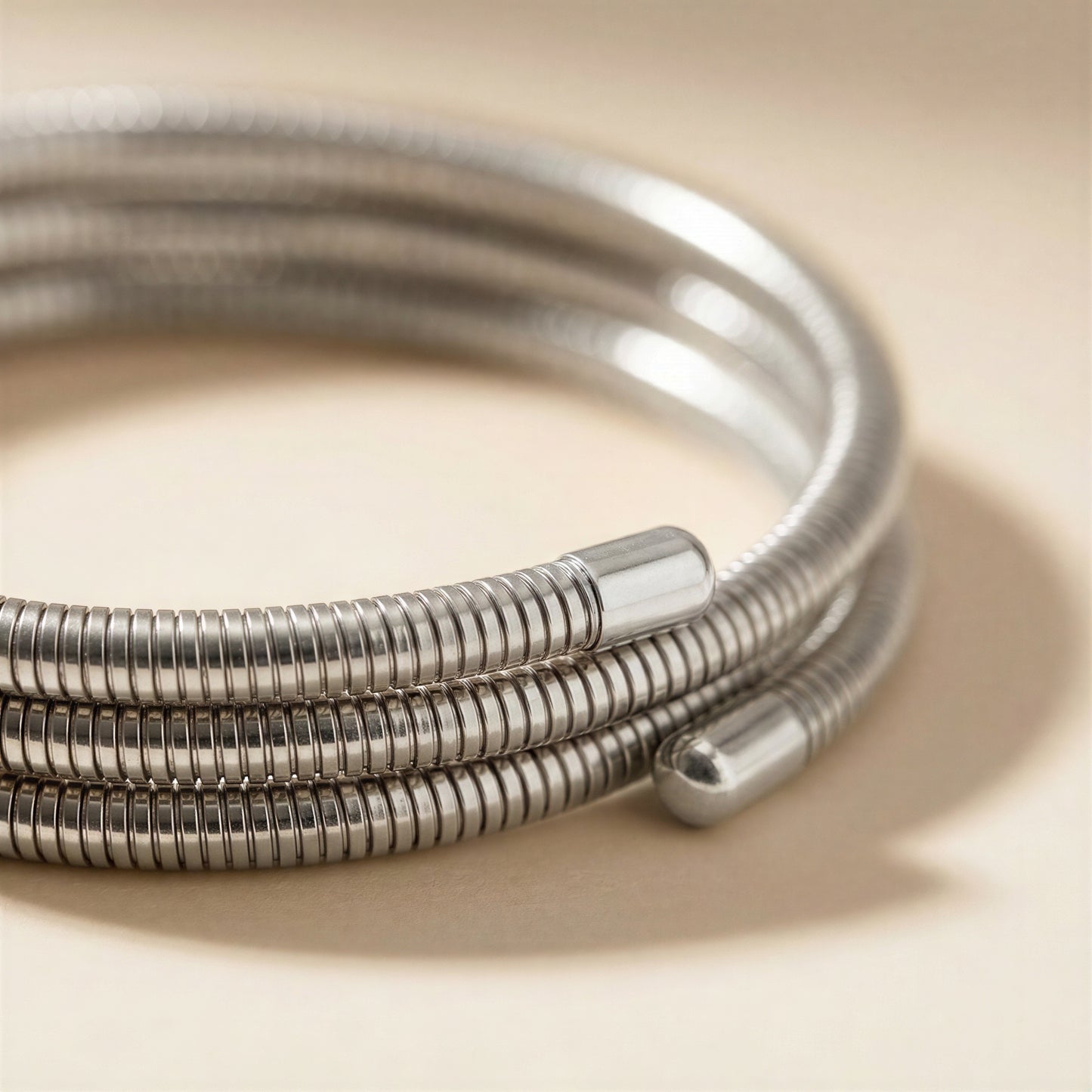 A close-up of a coiled silver metal bangle bracelet with a textured ridged outer surface and smooth cylindrical end caps, resting on a warm beige surface.