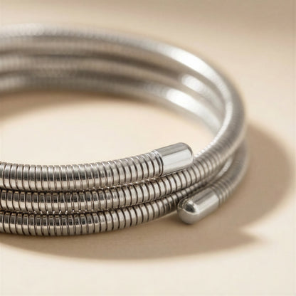A close-up of a coiled silver metal bangle bracelet with a textured ridged outer surface and smooth cylindrical end caps, resting on a warm beige surface.
