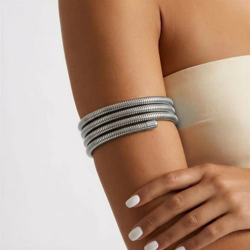A close-up of a person's upper arm and hand wearing a stack of four metallic silver coiled bangles against a light neutral background.