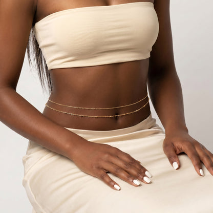 A close-up of a model wearing a delicate double-strand gold chain anklet on her ankle, styled with a matching beige bandeau top and skirt.