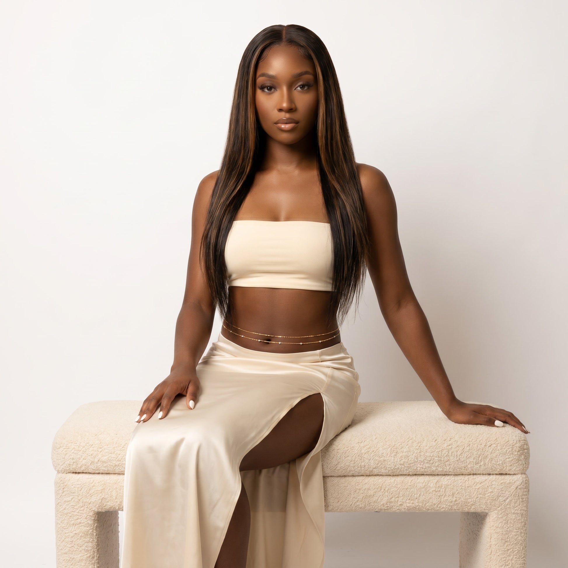 A woman with long, straight dark brown hair sits on a textured cream-colored bench against a plain off-white background. She is wearing a matching beige two-piece set consisting of a strapless tube top and a high-slit maxi skirt, accessorized with a thin gold chain waist belt.